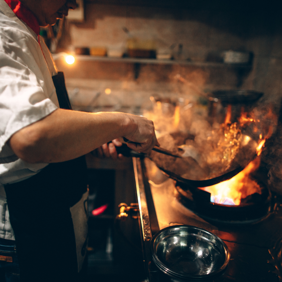 A chef in a white shirt and apron cooks over a high flame, stirring ingredients in a wok as if crafting a culinary journey to the Heart of Patagonia. The dimly lit kitchen, with its array of utensils and containers, highlights the vibrant cooking process.