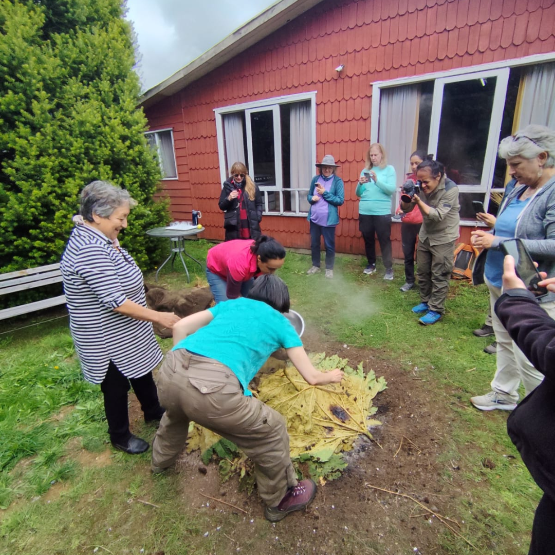 On a Private Trip in the Heart of Patagonia, a group watches as two women, one bending over and one standing, prepare food covered with large leaves outside a red wooden building. Steam rises from the feast while onlookers snap photos.