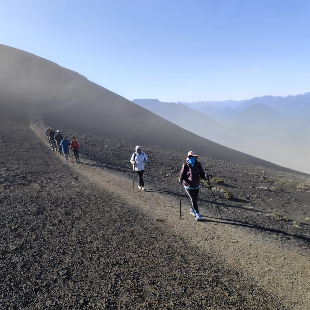 A group of hikers with trekking poles walk up a rocky, barren mountain trail under a clear blue sky, with distant mountains in the background—an unforgettable moment on a Patagonia private trip.