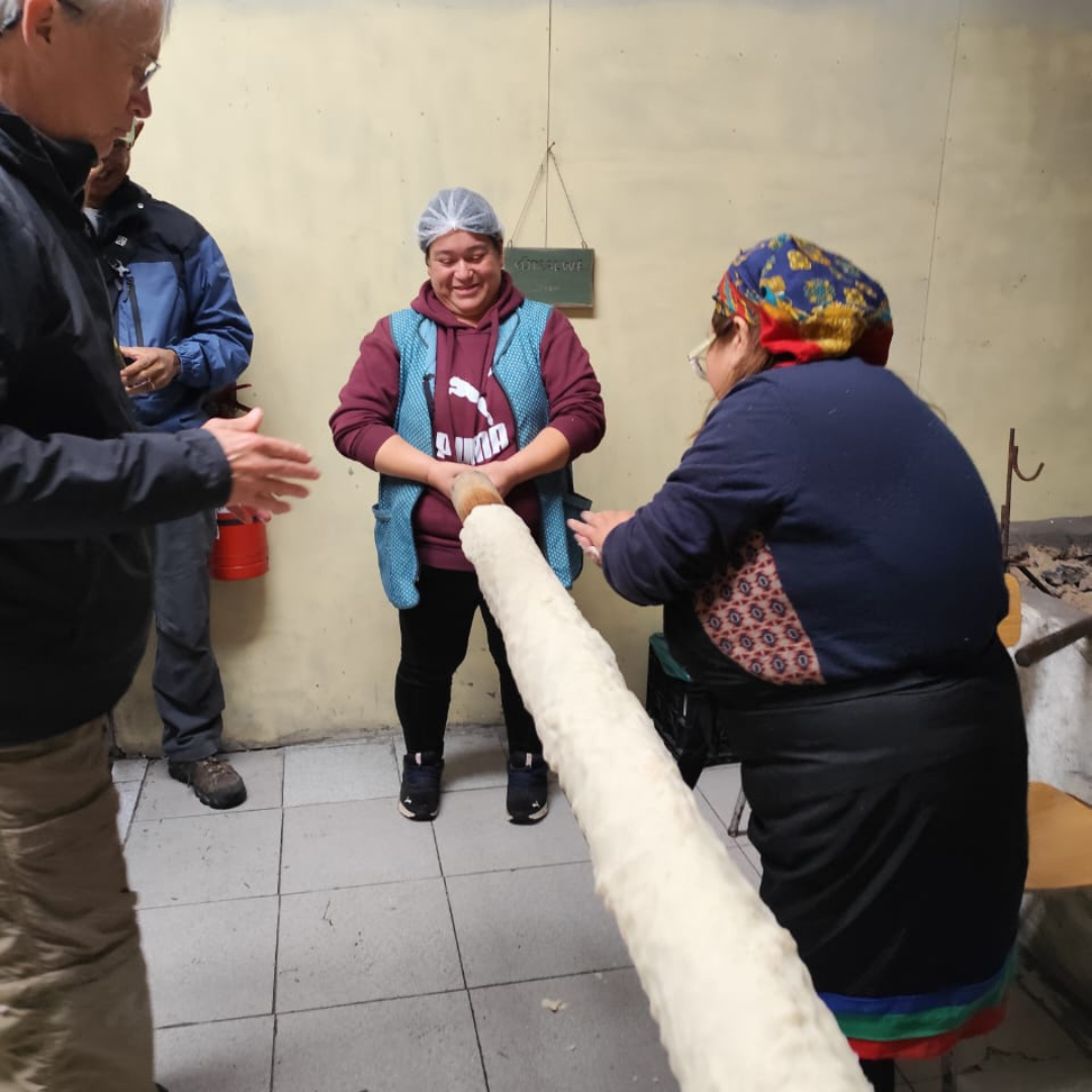 Three people stand indoors holding a long rolled piece of dough. The woman in front wears a colorful headscarf and apron, while the woman in the middle smiles, and a man on the left gestures—a warm moment during a private trip in the Heart of Patagonia.