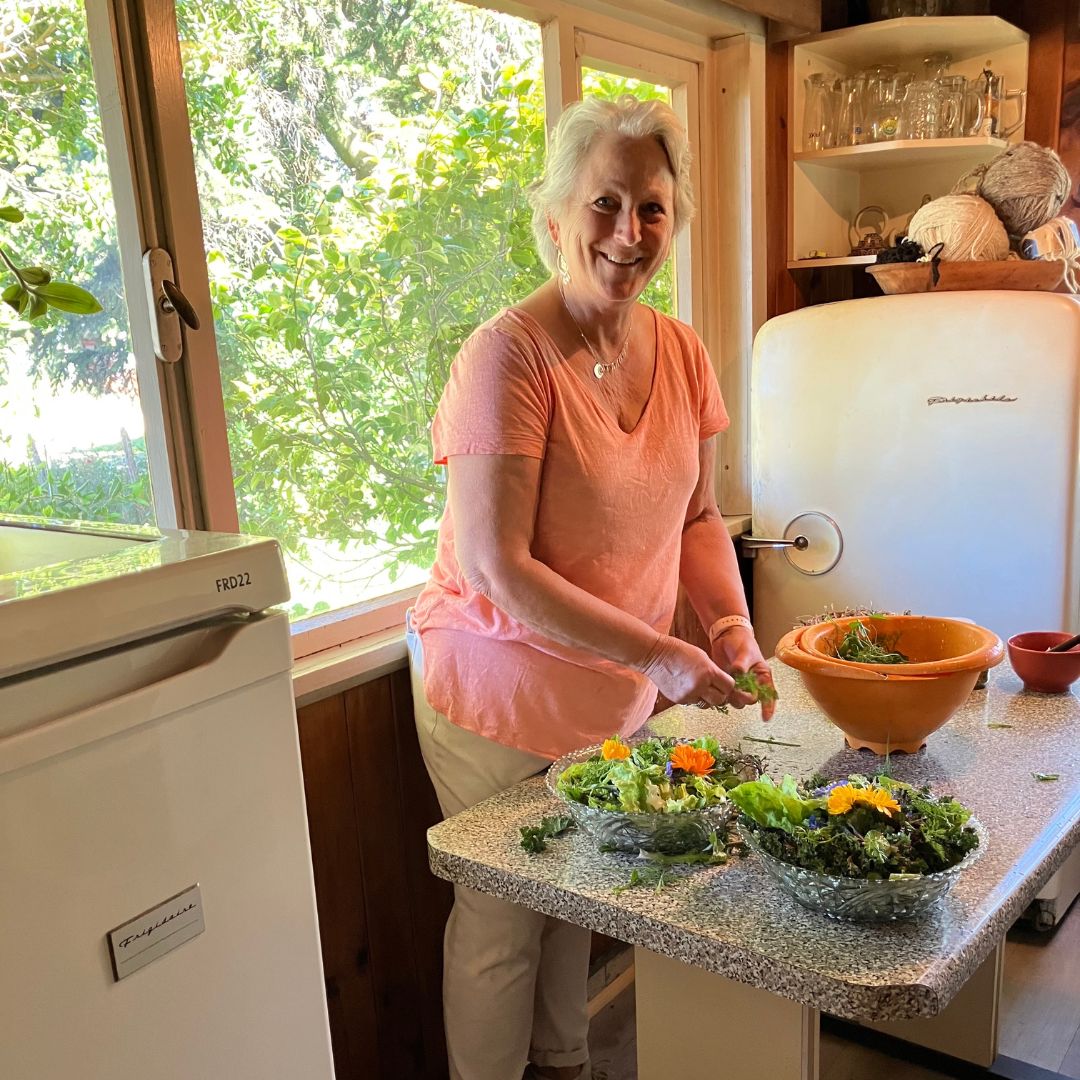An older woman smiles while preparing fresh salads at a kitchen counter beside a window, sunlight streaming in. Bowls of greens and a large orange mixing bowl await as travelers enjoy the cozy kitchen filled with vintage appliances.