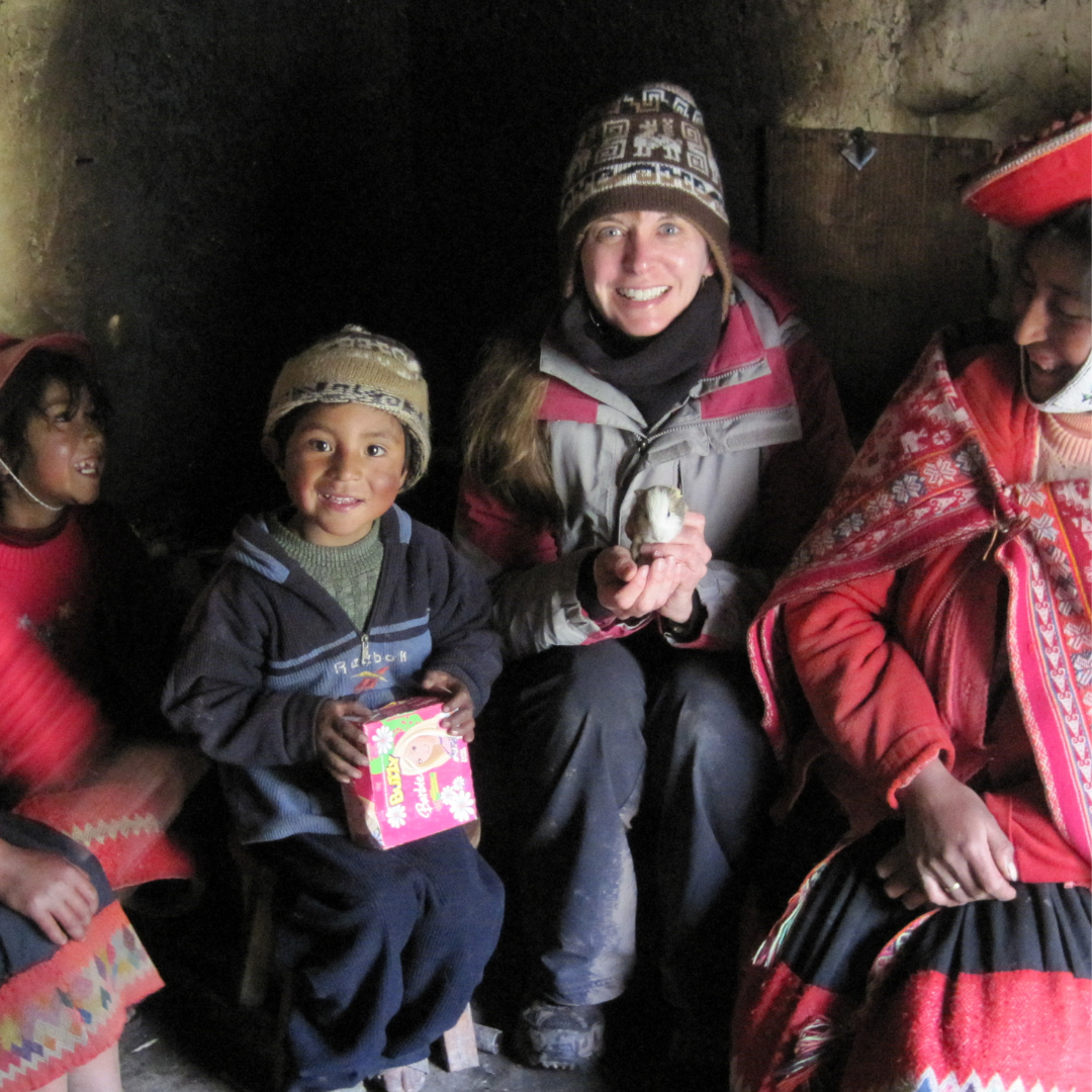 A woman in a winter hat and jacket smiles while holding a small white guinea pig, joining fellow travelers—including two children and a woman in traditional Andean clothing—inside a dimly lit room.