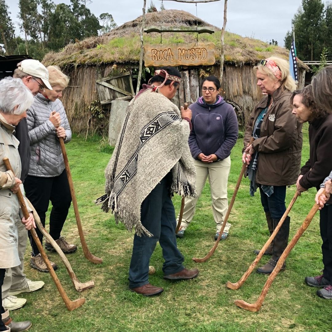 A group of travelers stand in a circle on grass, holding wooden staffs, listening to a man in a poncho and hat outside a rustic hut with a sign reading Ruca Museo. Trees and greenery are visible in the background.